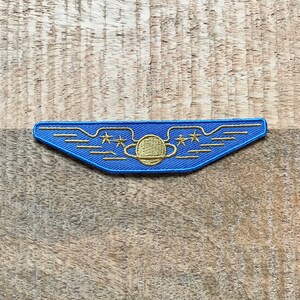 May include: A blue and gold embroidered patch with a planet and three stars inside a winged shape.