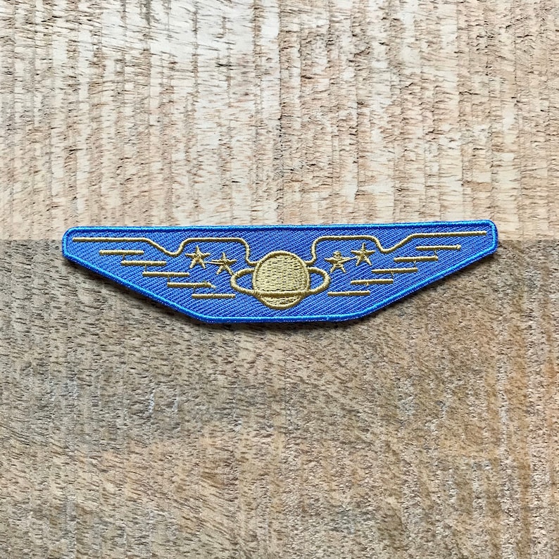 May include: A blue and gold embroidered patch with a planet and three stars inside a winged shape.