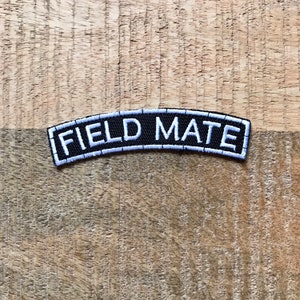 Puede incluir: Parche bordado en blanco y negro con el texto "FIELD MATE" en blanco sobre fondo negro.