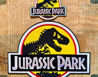 Könnte beinhalten: Zwei Jurassic Park-Patches. Der größere Patch zeigt eine schwarze Dinosaurier-Silhouette auf gelbem Hintergrund, mit rotem Rand und den Worten "JURASSIC PARK" in Weiß. Der kleinere Patch hat ein ähnliches Design. Beide Patches befinden sich auf einer Holzoberfläche.