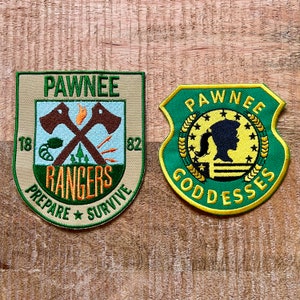 Peut inclure: Deux écussons brodés. Le premier est beige avec des accents verts et orange, représentant des haches croisées et le texte "Pawnee Rangers". Le second est vert avec des accents jaunes, affichant une silhouette et le texte "Pawnee Goddesses".
