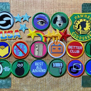 Peut inclure: Une collection de 16 patchs brodés colorés avec différents motifs, dont un pingouin, un hibou, un arc-en-ciel, des toilettes, une batterie, une empreinte de patte, une étoile, un drapeau 'USA' et le texte 'Pawnee Goddesses'.