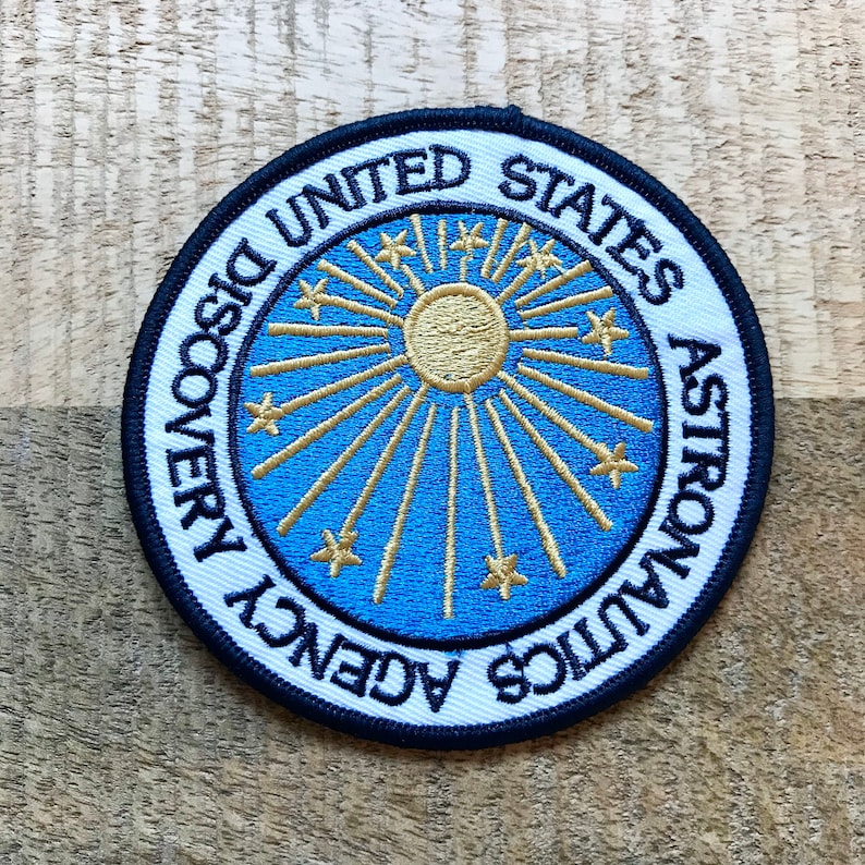 May include: A white, blue, and gold embroidered patch with the text "United States Astronauts Discovery Agency" in a circle. The center of the patch features a sun with rays and stars.