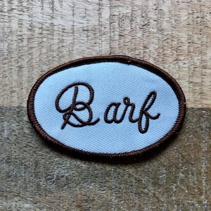 Peut inclure: Un écusson ovale blanc avec une bordure brodée marron. Le mot "Bark" est brodé en lettres cursives marron sur l'écusson.