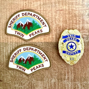 Könnte beinhalten: Zwei gestickte Aufnäher und ein goldfarbener Abzeichen. Die Aufnäher sind weiß mit dem Text "SHERIFF DEPARTMENT TWIN PEAKS" um eine Berg- und Waldszene. Das Abzeichen lautet "TWIN PEAKS DEPUTY 13" mit einem blauen Stern.