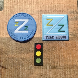 Könnte beinhalten: Drei gestickte Patches: ein blauer Kreis mit dem Text "Team Zissou Master Frogman" und einem weißen "Z" mit gelben und grünen "Z"s darin, ein hellblaues Quadrat mit einer weißen Diagonalen und gelben und grünen "Z"s und eine schwarze Ampel mit roten, gelben und grünen Lichtern.