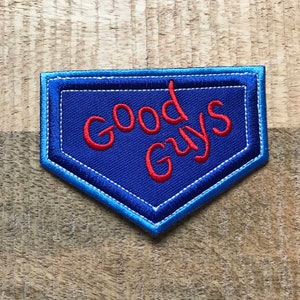 Può includere: Una toppa blu ricamata con le parole "Good Guys" in corsivo rosso. La toppa ha un bordo bianco e un contorno blu, su uno sfondo di legno. La toppa ha la forma di un piatto di casa base.