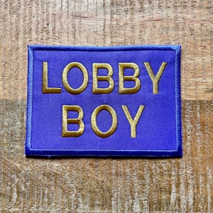 Może przedstawiać: Naszywka z niebieskiego materiału z haftowanym złotym tekstem "LOBBY BOY".