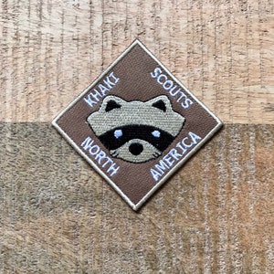 Può includere: Una toppa marrone a forma di rombo con il testo "Khaki Scouts North America" attorno ai bordi. Al centro della toppa c'è un viso di procione ricamato in bianco.