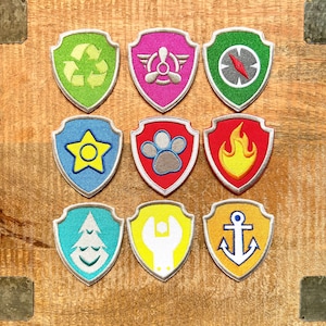 May include: Nine embroidered shield-shaped patches with various designs. These include a recycling symbol, propeller, compass, star, paw print, flame, tree, wrench, and anchor. The patches are in a range of colours: green, pink, blue, red, yellow, and teal. They are set on a wooden surface.