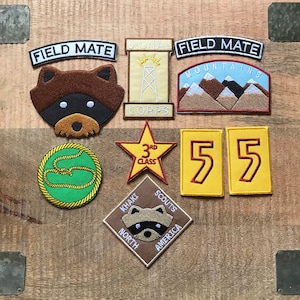 Peut inclure: Une collection de neuf patchs brodés sur le thème du scoutisme. Les patchs sont jaunes, bruns, verts et rouges avec du texte blanc. Les patchs comprennent "Field Mate", "Signal Corps", "Mountains", "3rd Class", "Khaki Scouts North America", et "55".