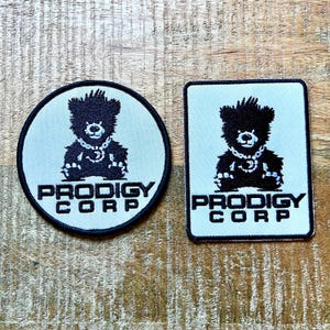 May include: Two embroidered patches, one circular and one rectangular, featuring a black bear graphic wearing a chain necklace. Both patches have a light-coloured background with a black border and the black text PRODIGY CORP printed below the bear.