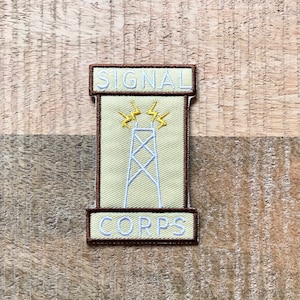Op de afbeelding: Een bruin en beige geborduurd patch met de tekst "SIGNAL CORPS" en een witte omtrek van een radiotoren met bliksemschichten bovenop.