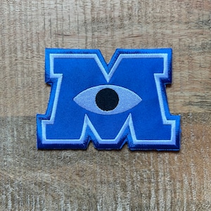 May include: Blue felt patch with a white outline in the shape of the letter M. A white eye with a black pupil is centred in the M.