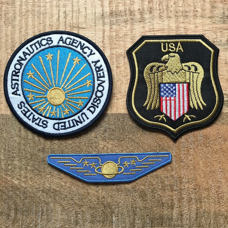 May include: Three embroidered patches, one circular with the text "United States Aeronautics Agency Discovery" and a sun with stars, one shield-shaped with the text "USA" and an eagle with an American flag, and one blue wing-shaped patch with a planet and stars.