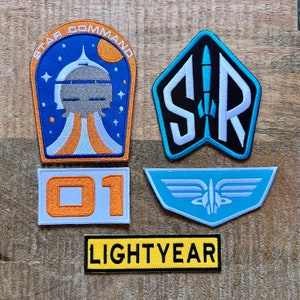 Può includere: Una collezione di toppe ricamate con disegni a tema spaziale. Include una toppa con il testo "STAR COMMAND", un razzo, il numero "01" e la parola "LIGHTYEAR". Le toppe sono in vari colori tra cui blu, arancione e bianco.