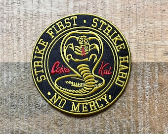 Puede incluir: Un parche bordado negro y dorado con una serpiente cobra y el texto "Strike First Strike Hard No Mercy" y "Cobra Kai".
