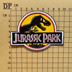 Op de afbeelding: Geborduurde patch met het "Jurassic Park" logo. Het heeft een zwart Tyrannosaurus Rex silhouet op een gele achtergrond, met "JURASSIC PARK" in wit en rood. De patch is ongeveer 10 cm lang. Een klassiek item met filmthema.