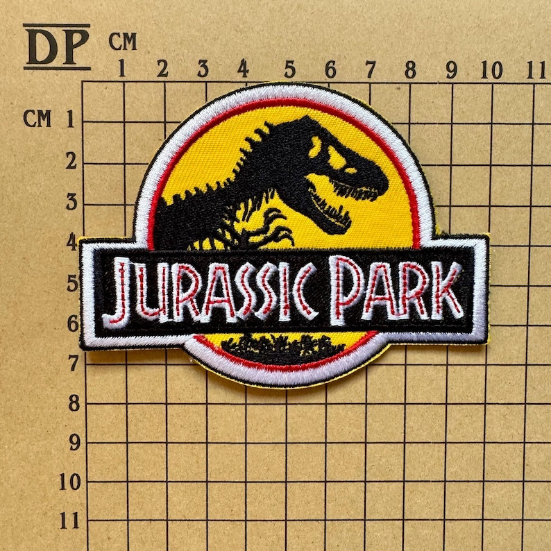 Op de afbeelding: Geborduurde patch met het "Jurassic Park" logo. Het heeft een zwart Tyrannosaurus Rex silhouet op een gele achtergrond, met "JURASSIC PARK" in wit en rood. De patch is ongeveer 10 cm lang. Een klassiek item met filmthema.