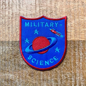 Puede incluir: Un parche bordado azul con un cohete rojo, un planeta rojo con un anillo azul y estrellas. El texto "MILITARY-SCIENCE" está bordado en azul en el parche.