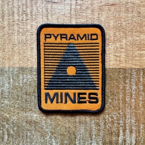 以下が含まれることがあります： オレンジと黒の刺繍パッチで、「PYRAMID MINES」というテキストと、白い円が内側に描かれた黒い三角形があります。