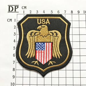 May include: A black and gold embroidered patch with a shield shape. The shield features a stylized American bald eagle with outstretched wings, holding a shield with the American flag. The text "USA" is above the eagle.