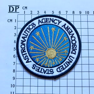 May include: A white, blue, and gold embroidered patch with the text "United States Astronautics Agency Discovery".
