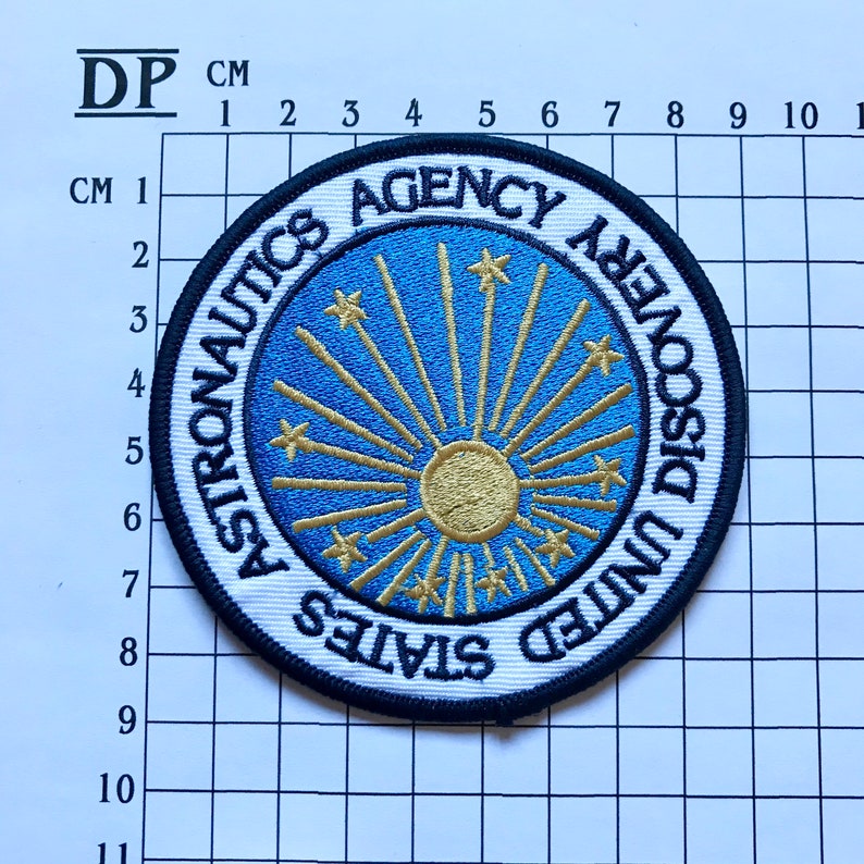 May include: A white, blue, and gold embroidered patch with the text "United States Astronautics Agency Discovery".