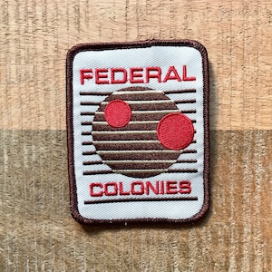 May include: A white embroidered patch with brown stitching. The patch features a brown circle with two red circles inside. The text "FEDERAL COLONIES" is embroidered in brown on the patch.