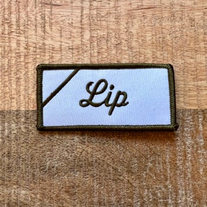 Puede incluir: Un parche de tela blanco con un borde verde y la palabra "Lip" bordada en letras cursivas negras.