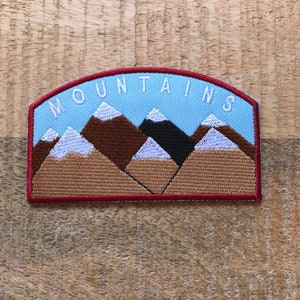 Può includere: Toppa ricamata con bordo rosso e sfondo blu. La toppa presenta una scritta bianca "MOUNTAINS" e una rappresentazione di cinque montagne con cime innevate bianche su uno sfondo di cielo blu.