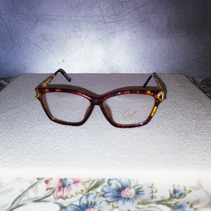 Puede incluir: Gafas de carey con montura de ojo de gato. Las gafas tienen detalles dorados en las patillas y un pequeño logotipo en la lente. La montura es de color marrón oscuro con un patrón de carey.