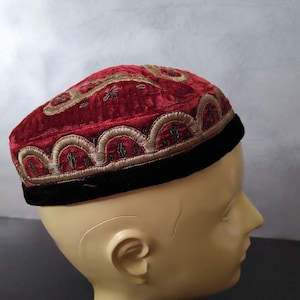 May include: A red velvet skullcap with gold embroidery. The skullcap has a black band around the bottom.