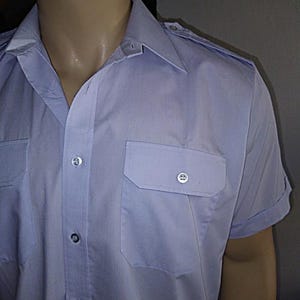 Puede incluir: Una camisa de manga corta azul claro con cierre de botones y dos bolsillos de solapa. La camisa tiene cuello y un solo botón en cada hombro.