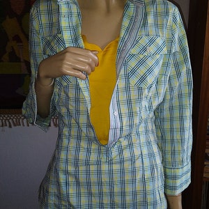 May include: A light blue and yellow plaid shirt with long sleeves and a button-down front. The shirt is unbuttoned and has a yellow undershirt visible.