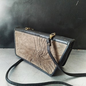 May include: A black leather clutch bag with a brown suede flap featuring a carved design. The bag has a long adjustable strap.