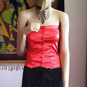 May include: A red satin strapless top with button details is paired with black pants. A black beaded necklace and a red hat complete the ensemble. The outfit is displayed on a mannequin.