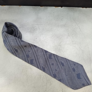 May include: A gray tie with a pattern of dark gray vertical stripes and abstract shapes. The tie is rolled up at the top and lies flat, showing its full length. The tie is made of a smooth fabric.