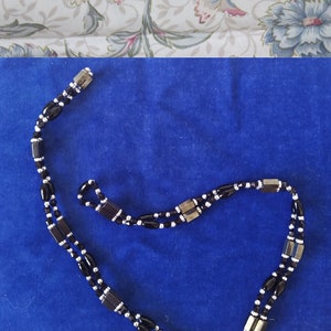 May include: A double strand necklace with black oval beads and silver spacer beads. The necklace has a silver clasp and is on a blue background.