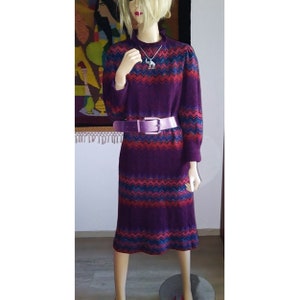 Könnte beinhalten: Ein vintage lila Kleid mit einem Zickzackmuster in Rot und Blau. Das Kleid hat lange Ärmel und einen hohen Ausschnitt. Ein hellvioletter Gürtel schnürt die Taille. Eine silberne Halskette mit Anhänger ist sichtbar.