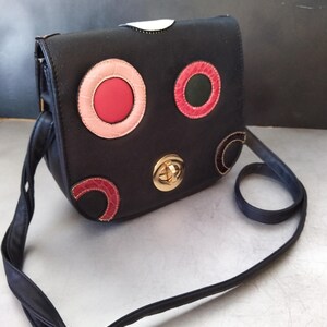 May include: A black crossbody bag with a gold clasp and colorful circle designs. The circles are made of different materials, including leather and fabric.