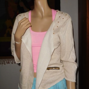 May include: A mannequin wearing a light beige blazer with gold-colored embellishments on the shoulders and a gold zipper pocket. Underneath, a pink tank top and turquoise shorts are visible. The outfit is casual and stylish.