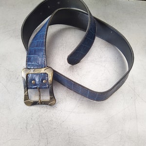 Peut inclure: Une ceinture bleue avec une texture imitation peau de crocodile. La ceinture a une boucle de couleur argentée avec un motif de feuille. La ceinture est faite d'un matériau bleu foncé et est incurvée en boucle.