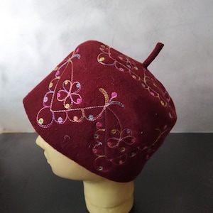 May include: A burgundy hat with colorful floral embroidery. The hat has a square crown and a small, upright stem. The embroidery features pink, yellow, blue, and green thread in a delicate floral pattern. The hat is displayed on a mannequin head.
