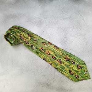 May include: A green necktie with a repeating geometric pattern in shades of yellow, orange, and black. The tie is rolled up at the top and lies diagonally across a light gray surface.