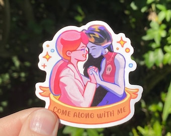 May include: A sticker featuring two cartoon characters, a pink-haired person with a white shirt and a blue-haired person with a black shirt, embracing. The sticker has a yellow banner with the text "Come along with me".