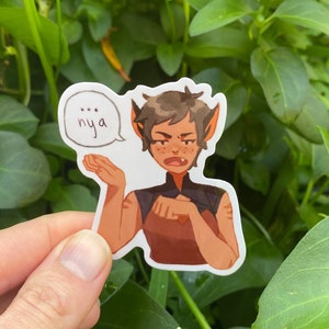 Op de afbeelding: Een sticker met een cartoonfiguur met bruin haar en groene ogen, die een bruin en gouden outfit draagt. Het figuur maakt een gebaar met zijn rechterhand en heeft een spraakballon boven zijn hoofd met de tekst "...nya".