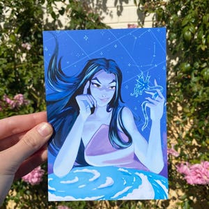 Peut inclure: Une impression d'art bleue représentant une illustration stylisée d'une femme aux longs cheveux foncés. La femme est représentée avec une expression pensive, sur un fond bleu avec des éléments célestes. L'œuvre a un thème fantastique.