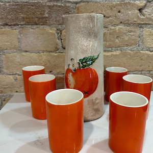 Puede incluir: Una jarra de cerámica vintage y seis vasos a juego. La jarra es beige con un diseño de manzana. Los vasos son de color naranja brillante con interiores blancos. El conjunto está dispuesto sobre una superficie blanca contra una pared de ladrillo.