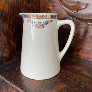 May include: White ceramic jug with a floral design in shades of pink, blue, and yellow. The jug has a wide mouth and a handle.